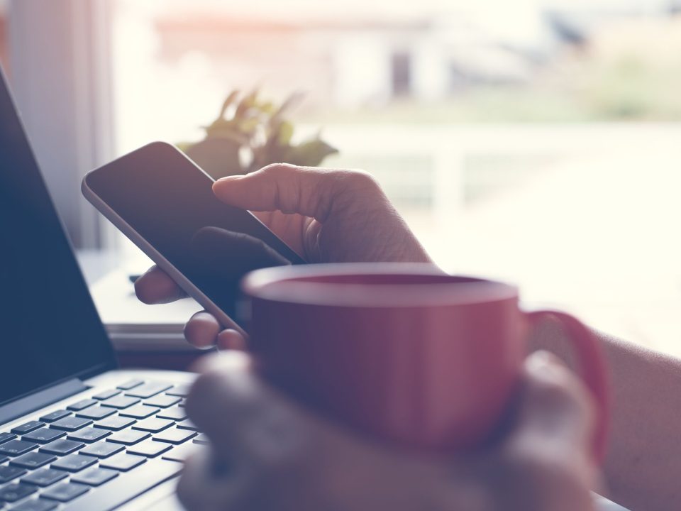 Person am Laptop mit Smartphone und Kaffeetasse in der Hand