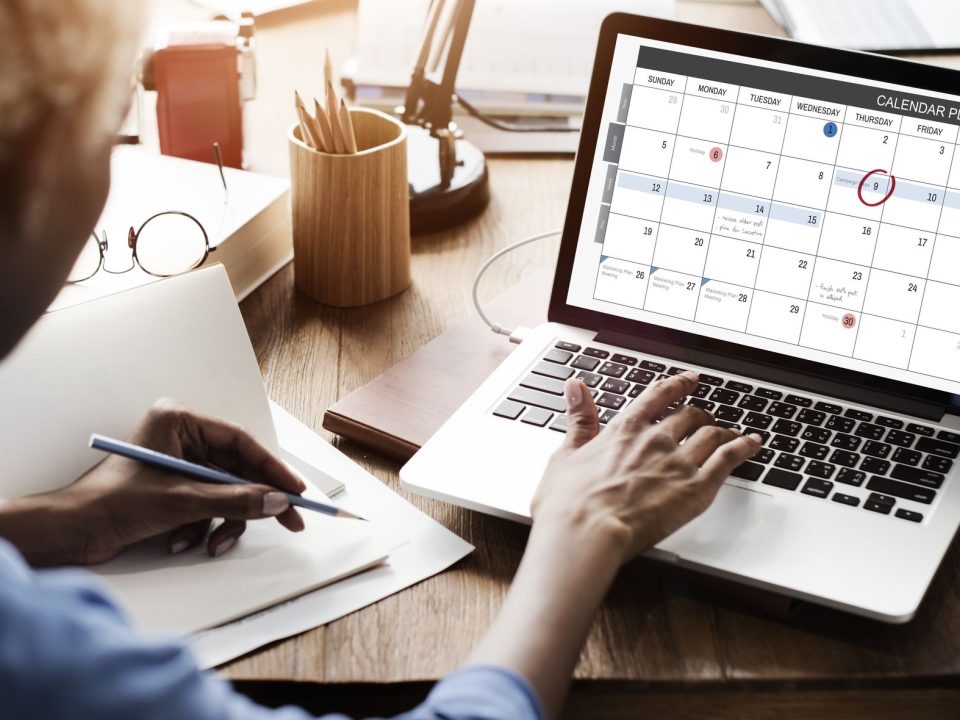 Person plant am Laptop mit geöffnetem Kalender und macht sich Notizen
